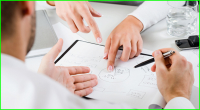 Two people pointing at a project plan on a desk.